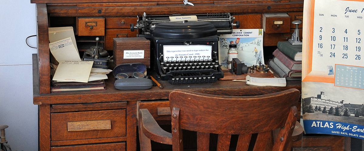 A close view of an antique wooden desk includes reading glasses, a telegraph machine, and a typewriter with a sign that reads: 