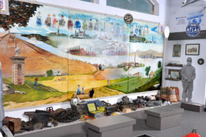 A mural at the Atlas Cement Memorial Museum includes depictions of a boat on a river, several factories, an ornamental post made of brick and concrete, grassy fields, the Statue of Liberty, and a row of figures in the blue sky representing Atlas employees. Below the mural are an array of artifacts including gearwheels, a stone block, and a Geiger counter. To the right is a not-quite-life-size cutout of a black-and-white photograph of a cement worker.