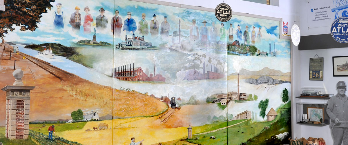 A mural at the Atlas Cement Memorial Museum includes depictions of a boat on a river, several factories, an ornamental post made of brick and concrete, grassy fields, the Statue of Liberty, and a row of figures in the blue sky representing Atlas employees. To the right is a not-quite-life-size cutout of a black-and-white photograph of a cement worker.