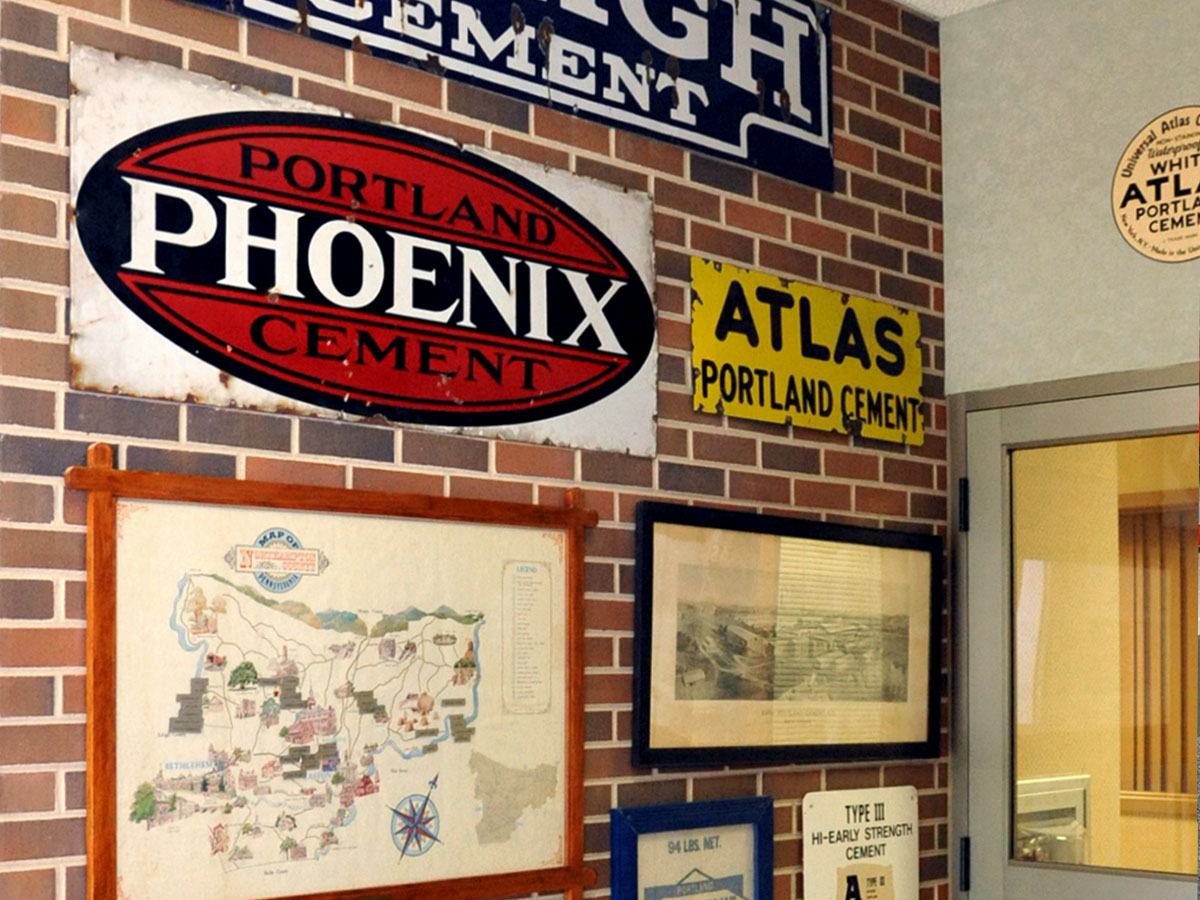 Metal signs, including ones for Phoenix Portland Cement and Atlas Portland Cement, are affixed to a brick wall along with historic maps and photographs.